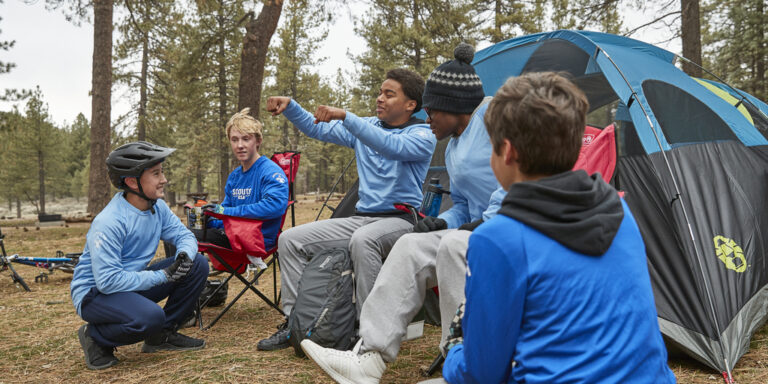 A group of Scouts exchange stories while seated in campchairs near a tent.